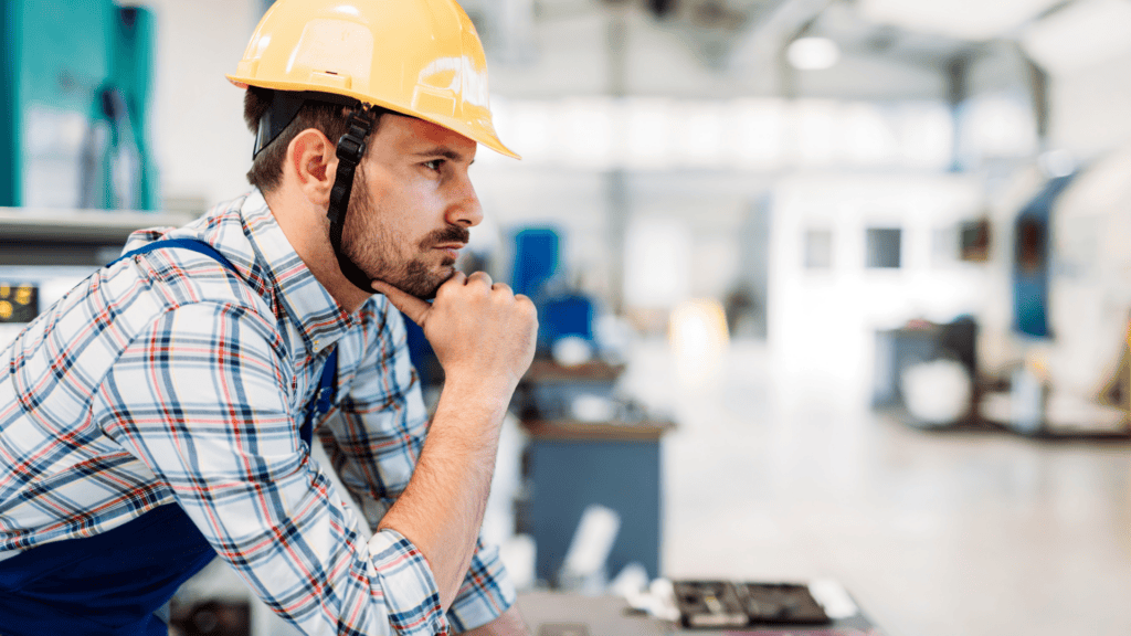 A man wearing a yellow hard hat and checkered shirt, with blue overalls, is leaning on a workbench, looking thoughtful. He is in an industrial setting, possibly a factory or workshop, with machinery and equipment in the background.
