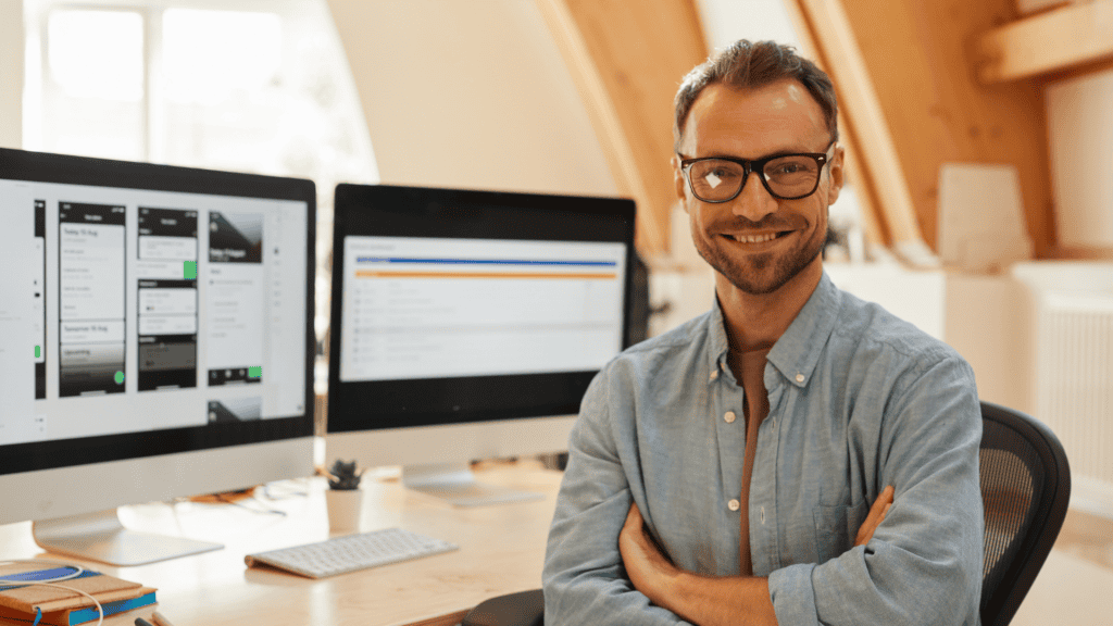 A man with glasses and a beard is smiling and sitting at a desk with his arms crossed. In the background, there are two computer monitors displaying different screens, one showing a design interface and the other displaying text or data, in a well-lit office space.
