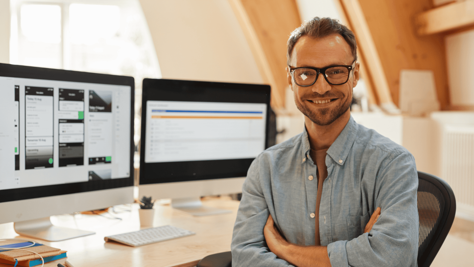 A man with glasses and a beard is smiling and sitting at a desk with his arms crossed. In the background, there are two computer monitors displaying different screens, one showing a design interface and the other displaying text or data, in a well-lit office space.