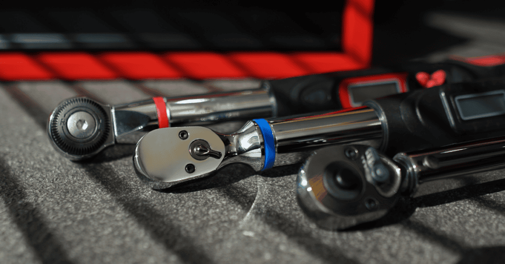 A close-up image of three metal ratchet wrenches laid out on a gray surface. Each wrench has a colored ring near the head: red, blue, and black. The wrenches have sleek, polished finishes, and their heads are angled slightly differently.