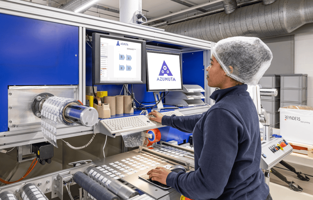 A worker, wearing a hairnet, operates a labeling machine in a manufacturing facility. The machine features a digital display screen showing branding for Azulmuta and is surrounded by various packaging materials. Rolls of labels are visible on the left side of the machine.
