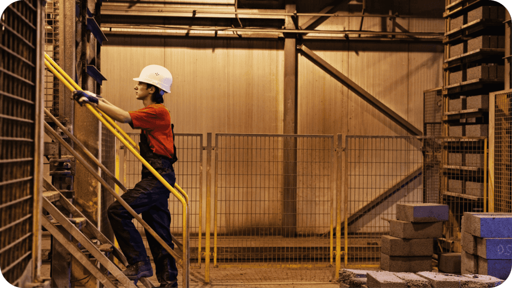 A construction worker wearing a white hard hat, red shirt, and dark overalls is climbing a yellow metal staircase in an industrial setting with concrete blocks and metal fencing in the background. The area is well-lit but appears to be indoor industrial.