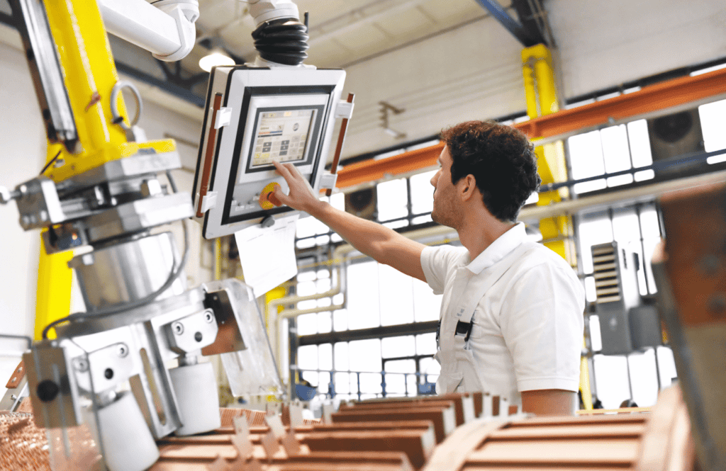 A man in a white shirt operates a touchscreen control panel in an industrial setting. The surrounding machinery and equipment suggest a manufacturing or production environment. The background shows large windows and structural elements of the facility.