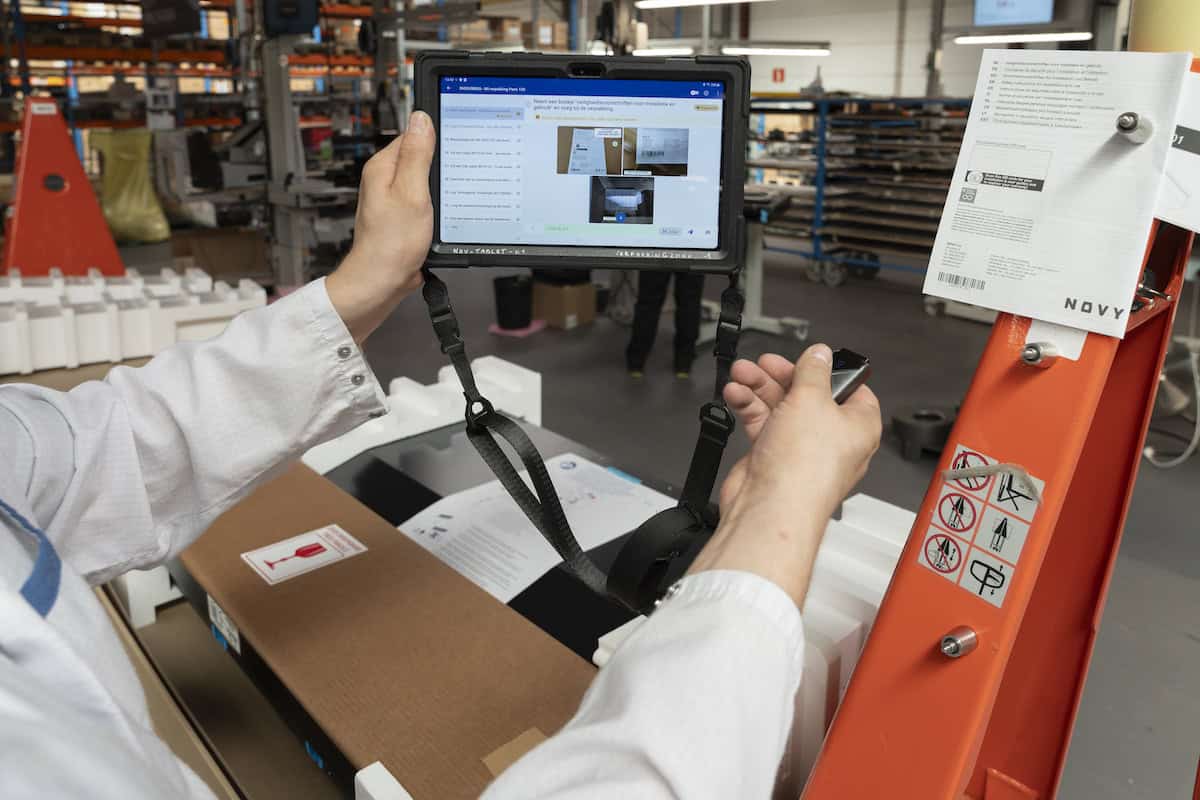 Een persoon in een labjas gebruikt een tablet om een assemblagechecklist te inspecteren in een productie- of industriële omgeving. Het tabletscherm toont afbeeldingen en tekst. Vlakbij zijn assemblageonderdelen, verpakkingsmaterialen en een machine met veiligheidsinstructies en labels te zien.