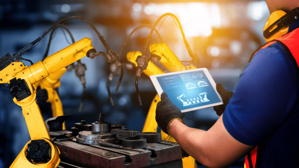 A worker in a blue shirt and orange safety vest uses a tablet to monitor or control yellow robotic arms in an industrial setting. The robotic arms appear to be working on a mechanical assembly, with a blurred manufacturing background.