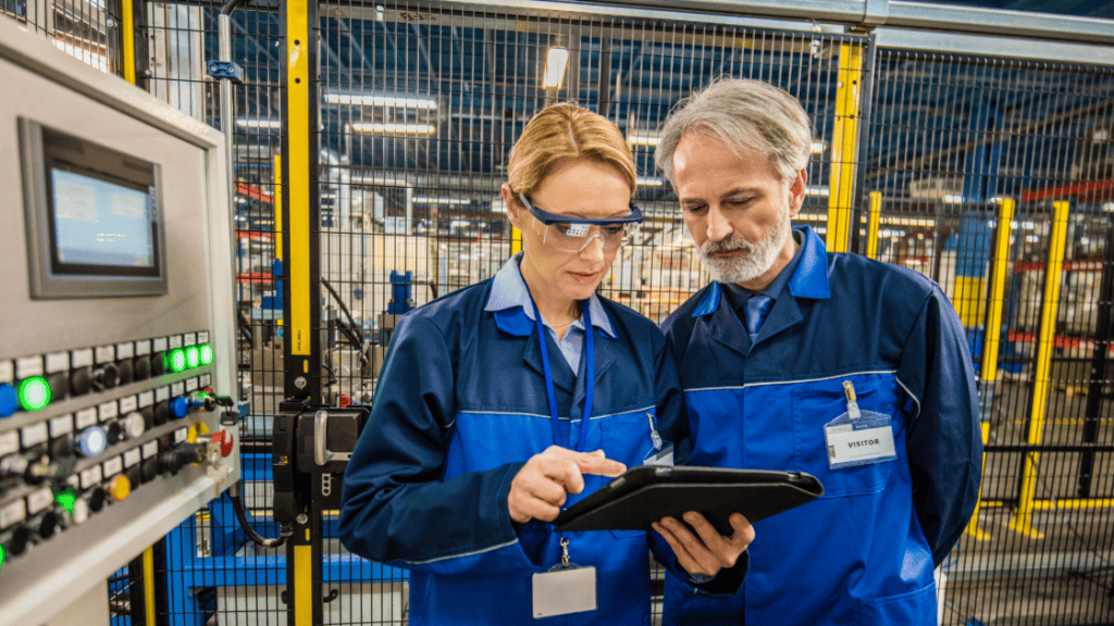 Two people in blue work uniforms, one wearing safety goggles, stand in a factory setting, closely examining a tablet. Behind them, there is industrial equipment and a cage-like structure. Control panels with illuminated buttons are visible to the left.