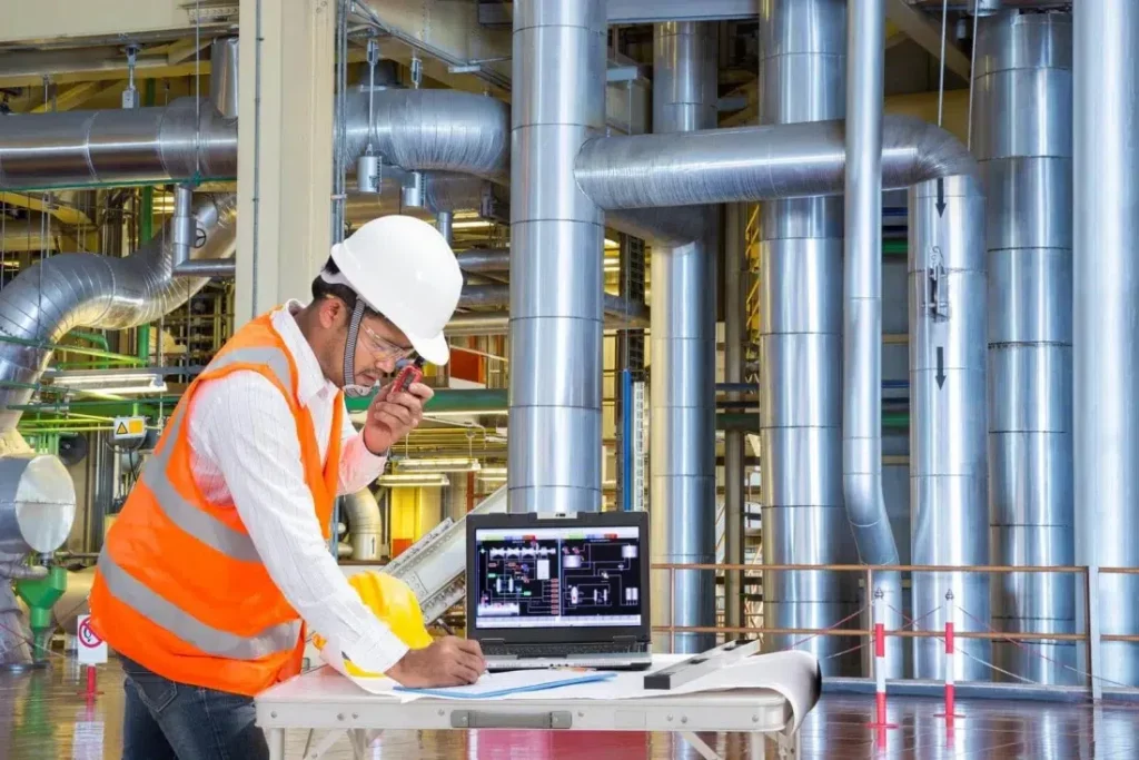 A construction worker in a high-visibility orange vest and white hard hat speaks into a walkie-talkie while writing on a clipboard. He stands near a laptop with industrial pipes and machinery in the background, suggesting an engineering or monitoring task.