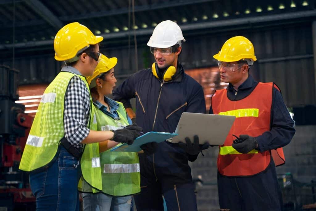 Four factory workers wearing safety gear, including hard hats, reflective vests, and safety goggles, stand together in a discussion. One worker holds a clipboard, while another holds a laptop. They appear to be collaborating in an industrial setting.