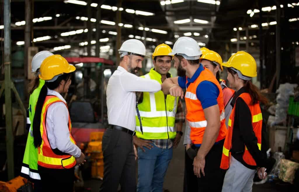 A group of construction workers, wearing protective helmets and reflective vests, stand together inside a warehouse. Two men in the center, one in a white shirt and the other in an orange vest, are bumping elbows. Others are observing and conversing.
