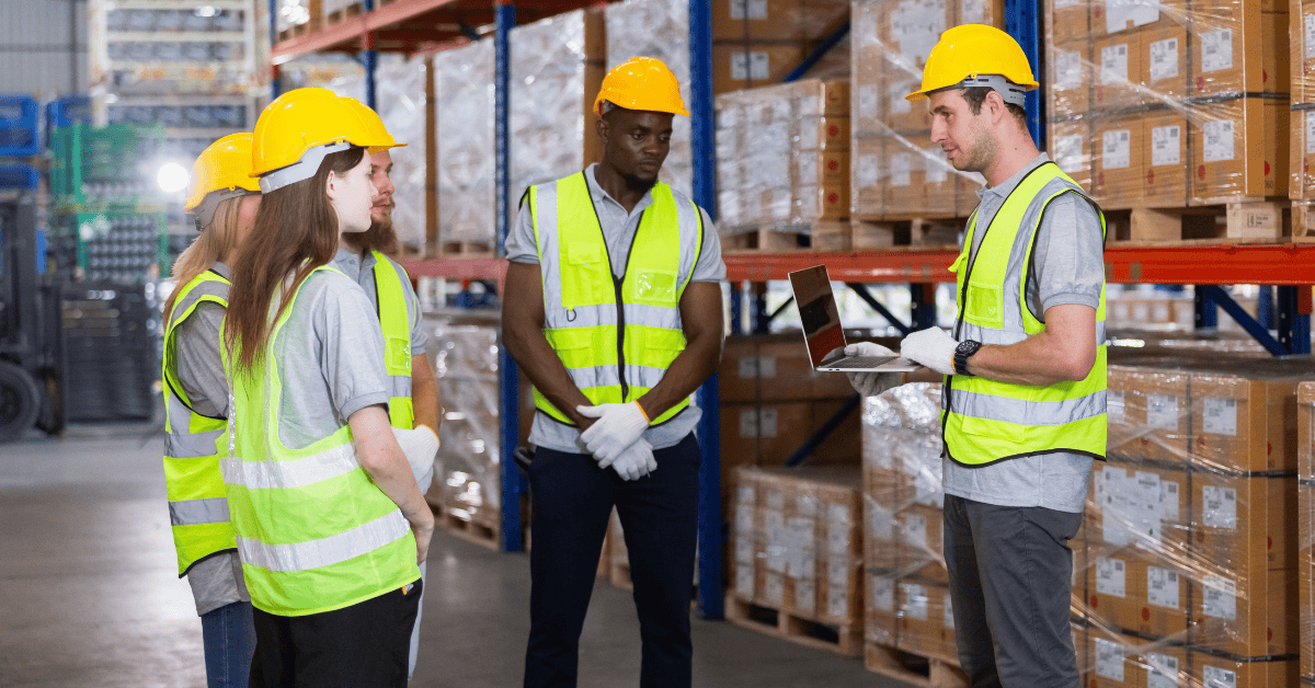 A group of five warehouse workers wearing high-visibility vests and yellow hard hats is listening to a male team leader with a laptop, who is also wearing a vest and hard hat. They are standing in a large warehouse with shelves stocked with boxes in the background.