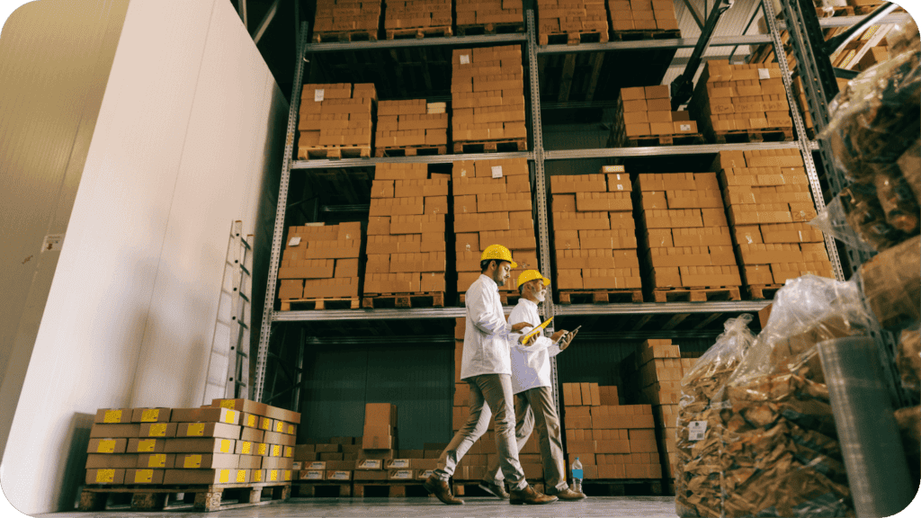 Two warehouse workers wearing yellow hard hats and white lab coats walk past tall shelves filled with stacked cardboard boxes. One holds a clipboard, and they appear to be inspecting inventory. The warehouse is well-organized and spacious.