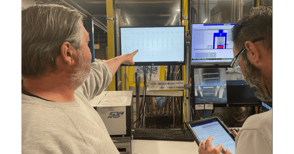 Two men are working in a technical environment. One is pointing to a large computer screen displaying data, while the other is using a tablet. There are multiple computer monitors and equipment in the background, indicating a high-tech workspace.