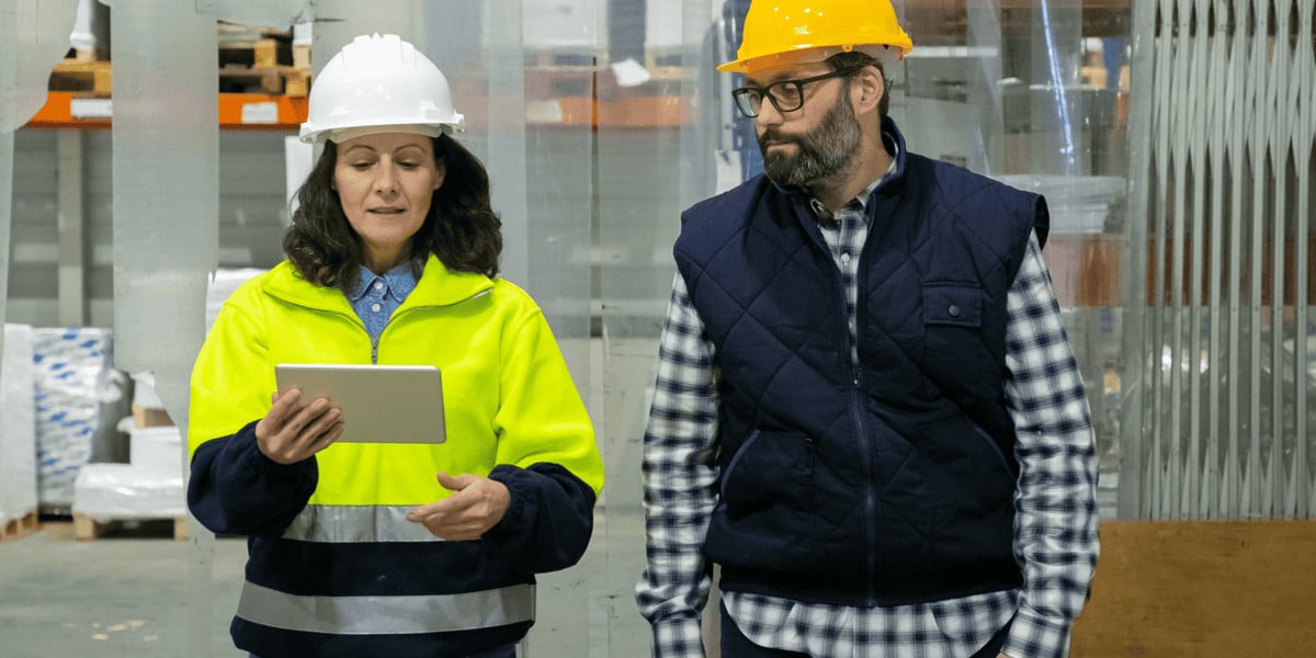 A woman wearing a high-visibility jacket and white hard hat instructs a man in a padded vest, flannel shirt, and yellow hard hat using a tablet computer in an industrial warehouse setting.