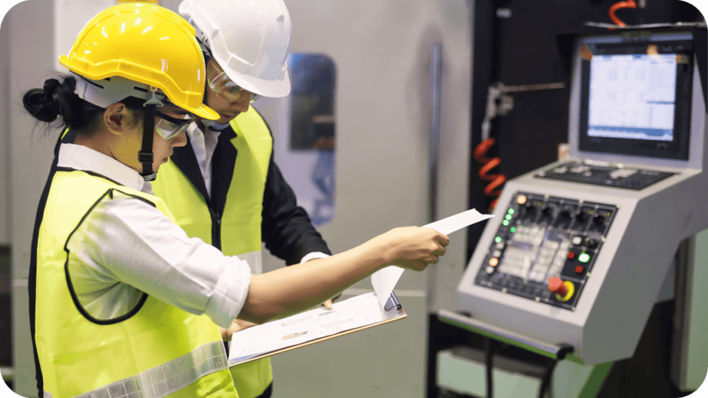 Two people wearing safety gear are working at a control panel in a manufacturing or industrial environment. One person points at the control panel while the other holds a clipboard. Both are wearing hard hats and high-visibility vests.