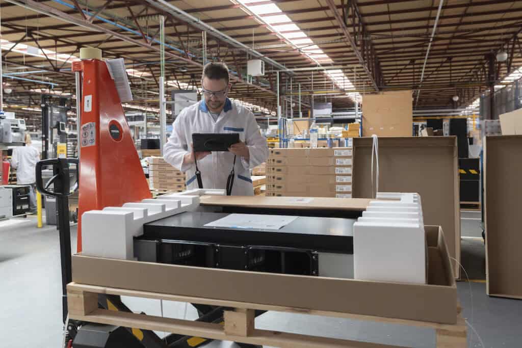 A worker in a white lab coat stands in a factory, using a tablet to inspect a partially assembled piece of equipment. The background shows a spacious industrial warehouse with shelves and various machinery. Packaging materials are visible around the equipment.
