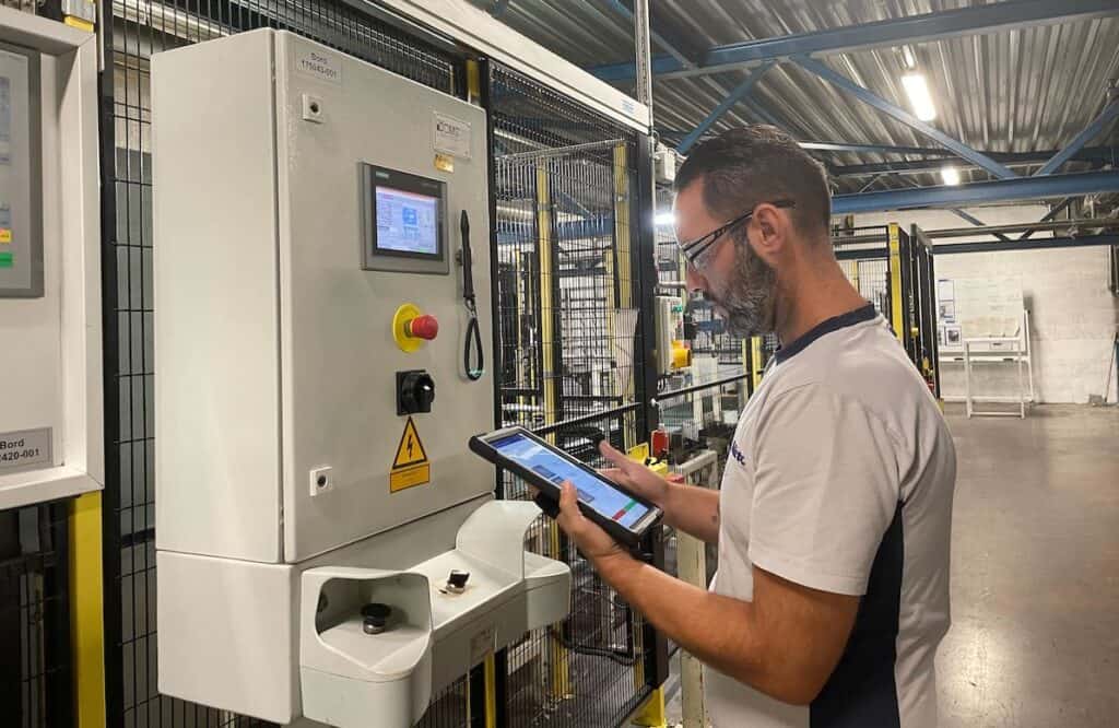 A man stands in a factory setting using a tablet device. He appears to be inspecting or programming an industrial control panel mounted on a wall. The factory features a steel structural framework and various machinery parts in the background.