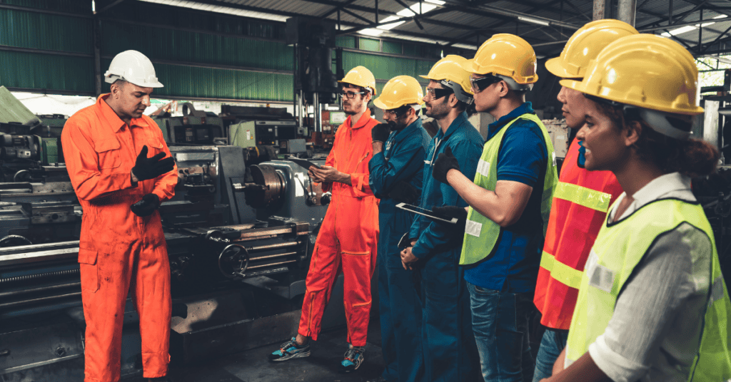 A group of factory workers, wearing safety helmets and protective clothing, listen attentively to a man in an orange jumpsuit explaining something. They are gathered indoors, surrounded by industrial machinery, in what appears to be a training session.