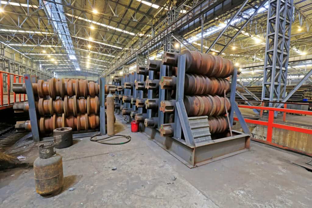 A large factory interior with metal beams and high ceilings. The space contains stacks of metal parts and large industrial cylinders. The floor has various equipment and gas cylinders scattered around, with overhead lights illuminating the area.