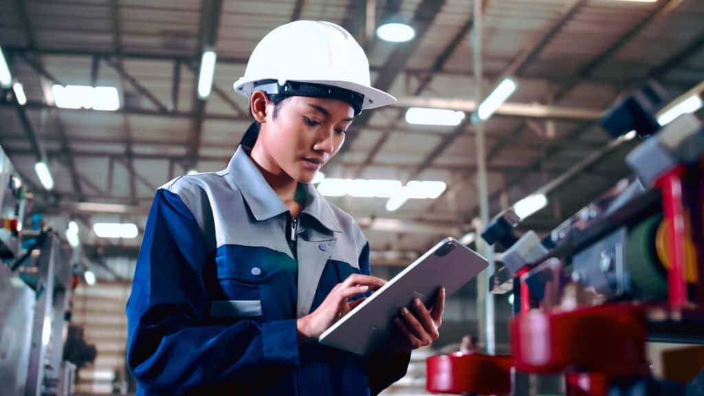 A worker wearing a hard hat and protective work attire stands in a factory setting, focused on a tablet device. The background features industrial equipment and machinery, with a high ceiling and metal beams visible overhead.