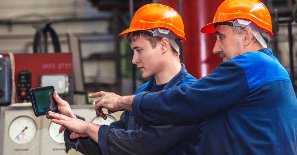 Two workers wearing orange safety helmets are in an industrial setting. The younger worker holds a tablet while the older worker points at it, seemingly instructing or discussing. Background includes industrial equipment with gauges and machinery.