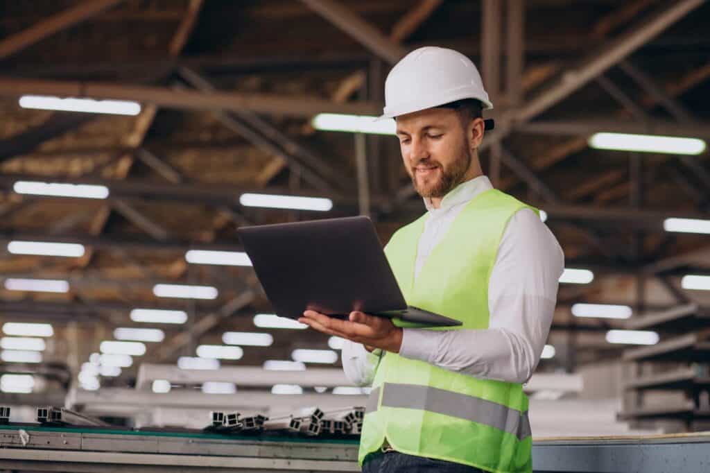 A man wearing a white hard hat and neon green safety vest stands in a warehouse. He is holding and typing on a laptop. The background shows industrial shelves and fluorescent lights.