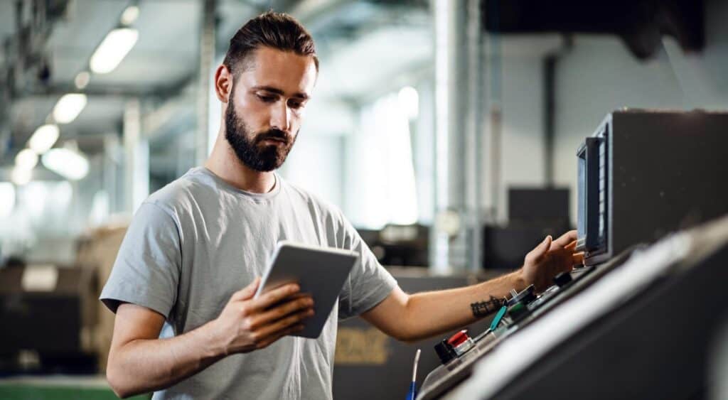 A man with a beard and short hair is standing in an industrial setting. He is focused on a control panel with one hand on the controls and holding a tablet in the other hand, likely adjusting or monitoring machinery. He is wearing a light gray T-shirt.
