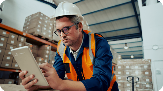 A warehouse worker wearing glasses, a white hard hat, and an orange safety vest is using a tablet. The background shows shelves filled with boxes and a high ceiling with lights.