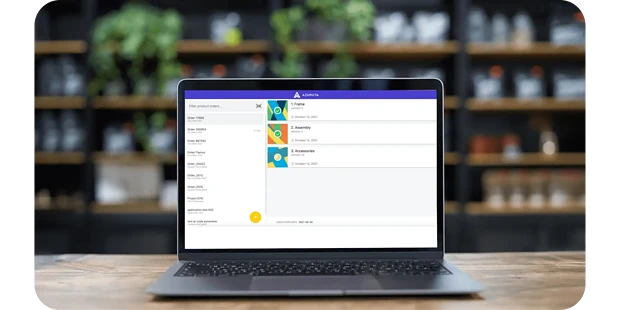 A laptop on a wooden table displays a colorful project management application on the screen with a purple banner at the top. The background shows blurred shelves adorned with plants and various items.