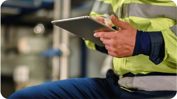 A person in a yellow high-visibility jacket and blue pants is holding a tablet with both hands. The background appears to be an industrial or work setting, with blurred machinery and equipment. Only the torso and hands of the person are visible.