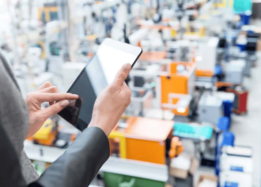 A person in business attire is holding and using a tablet, with a blurred background of a modern, automated industrial facility featuring various machinery and equipment.