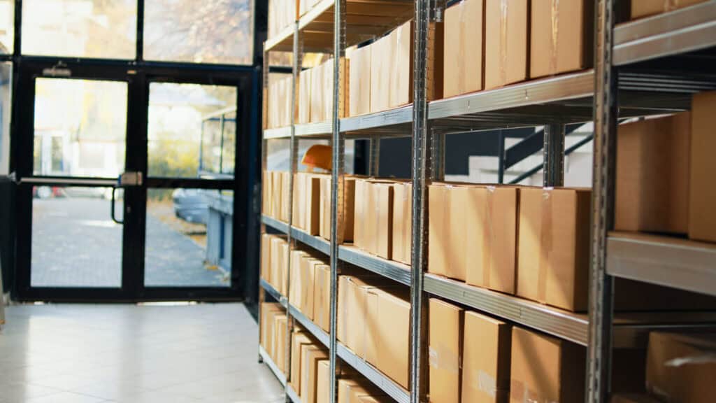 A storage room with metal shelves containing numerous brown cardboard boxes. The shelves are neatly organized and extend from the foreground to the background. Sunlight filters through glass doors at the end of the room, illuminating the space.