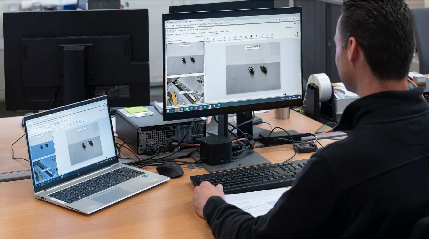 A person sits at a desk working on a computer with dual monitors and a laptop. One monitor displays an image editing software showing a close-up of small objects. The workspace includes various office equipment and papers.