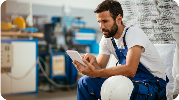 A man in a white t-shirt and blue overalls sits in a factory, holding a tablet. He appears to be reading or working on the device. On his lap is a white hard hat, and industrial equipment is visible in the background.