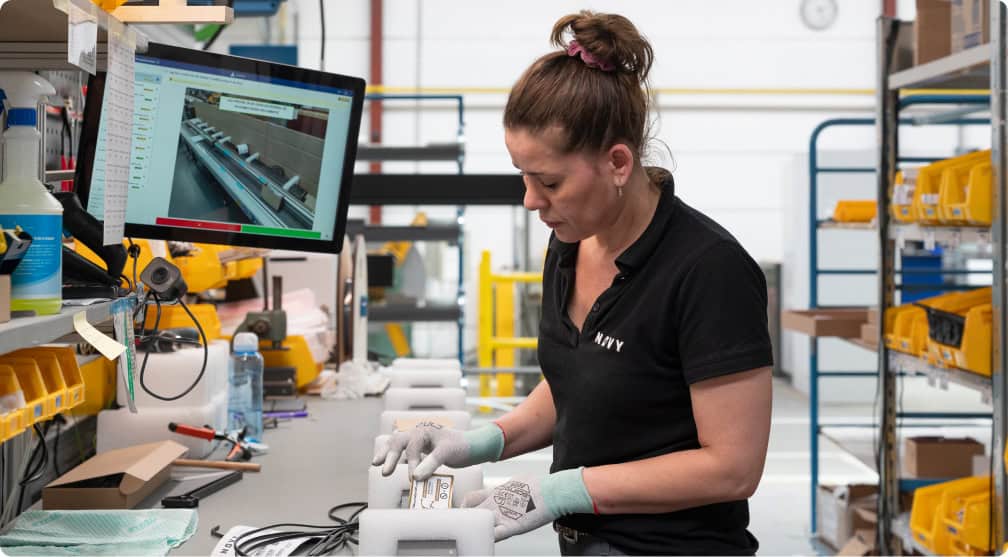 A woman wearing a black shirt with a company logo and gloves is standing at a workbench in an industrial setting. She is handling components and looking down at a device. A computer is on the left side of the bench, displaying a software interface.