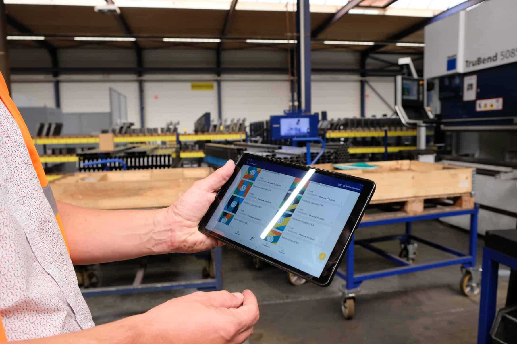 A person holds a tablet displaying industrial software in a manufacturing facility. In the background, there is machinery and equipment, suggesting an industrial or factory setting. The environment is well-lit with a focus on modern manufacturing technology.