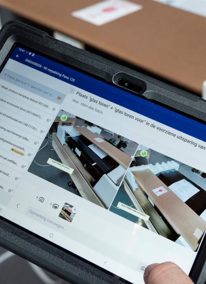 A person is holding a tablet that displays instructions for assembling or handling a piece of equipment. The screen shows two images with diagrams and text in Dutch related to placing 