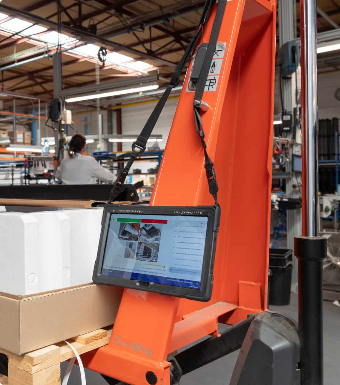 A tablet, attached to an orange industrial machine with black straps, displays information and images. The machine stands in a warehouse environment with cardboard boxes, white foam blocks, and other equipment in the background. A person is working in the distance.