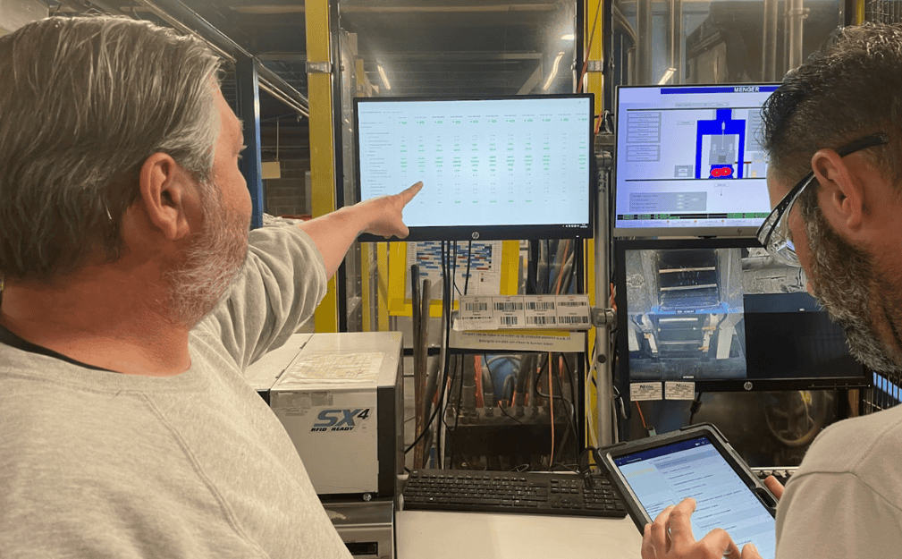 Two men are in a factory setting. One man with gray hair and a beard points to a large monitor displaying data, while the other man with dark hair and glasses looks at a tablet. Equipment and machinery are visible in the background.