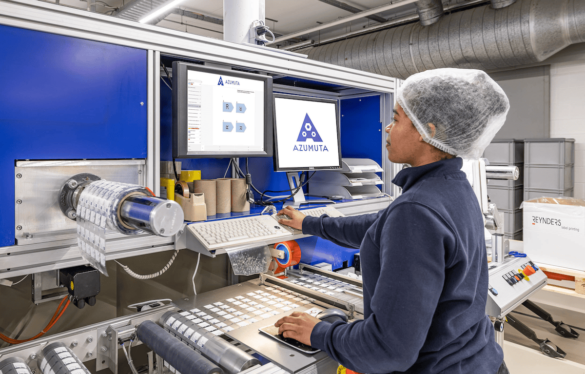 A worker operating a machine at a manufacturing facility. The machine displays the Azumuta logo on multiple screens. The worker is wearing a hairnet and dark clothing, interacting with the machine's keyboard and controls. Rolls of material are visible nearby.
