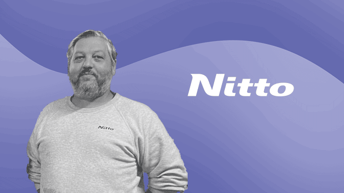 A man with a beard and short hair is wearing a light-colored sweatshirt with the word "Nitto" on it. He stands against a purple wavy background with the word "Nitto" prominently displayed in white.