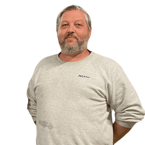 A man with gray hair and a beard is standing with his arms behind his back, wearing a light gray sweatshirt that has the word 