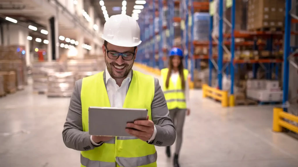 A smiling man wearing a white hard hat, glasses, and a neon yellow safety vest holds a tablet in a warehouse. In the background, a woman in a blue hard hat and safety vest walks towards him. Shelves filled with boxes and supplies line the walls.
