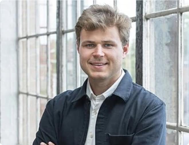 A man with short, wavy hair and a light beard stands in front of large, industrial-style windows. He is wearing a dark jacket over a white shirt and has his arms crossed, smiling at the camera. The background is bright and airy..