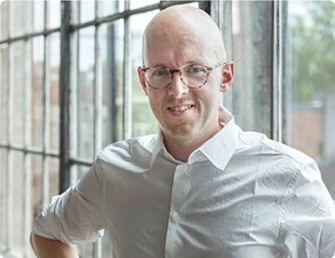 A person with glasses, wearing a white shirt, stands in front of large, industrial-style windows. The person has a bald head and is smiling slightly. The background shows a blurred outdoors, suggesting it is daytime.