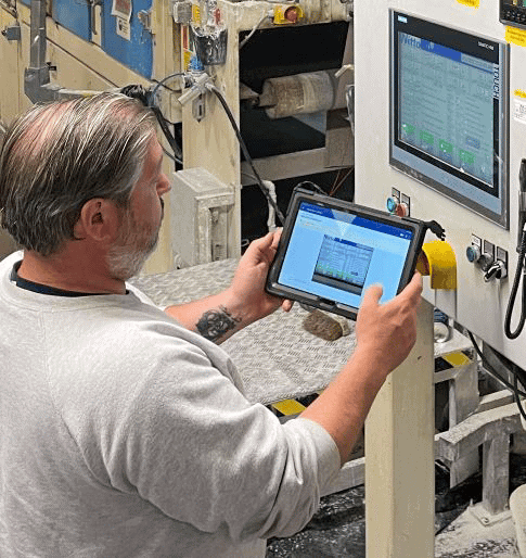 A man in a grey sweatshirt, holding a tablet, stands in front of a control panel with a touchscreen display. He appears to be monitoring or adjusting settings at an industrial or manufacturing facility. Machinery and equipment are visible in the background.