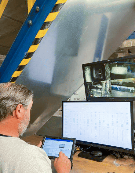 A person with grey hair operates a tablet in front of two monitors. One monitor shows surveillance footage, while the other displays a spreadsheet. The workstation is set against a backdrop of industrial equipment with yellow and black caution markings.