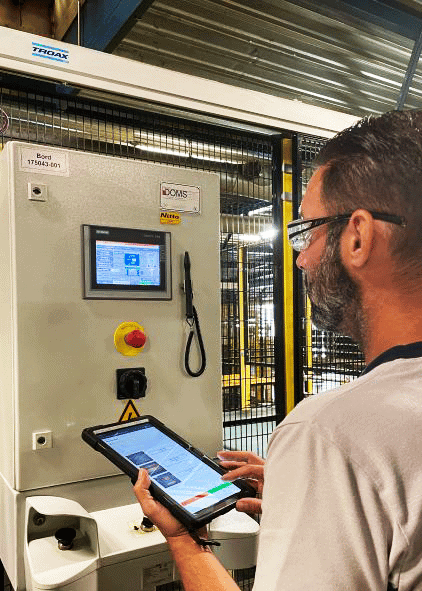 A man with glasses operates industrial machinery using a tablet and a control panel. The control panel features a screen displaying technical information, buttons, and emergency stop. The background shows industrial equipment with yellow-and-black safety barriers.