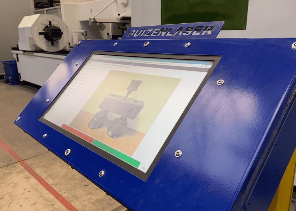 A blue industrial machine with a touchscreen display shows an image of a mechanical part on the screen. The words 