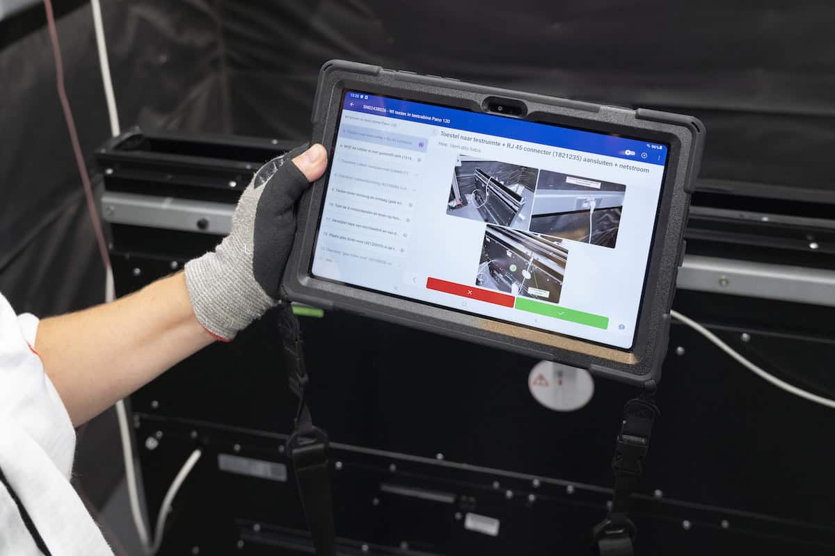 A person wearing a glove holds a tablet displaying technical instructions and images, possibly for machinery or equipment maintenance. The background shows industrial equipment in a workshop or factory setting.