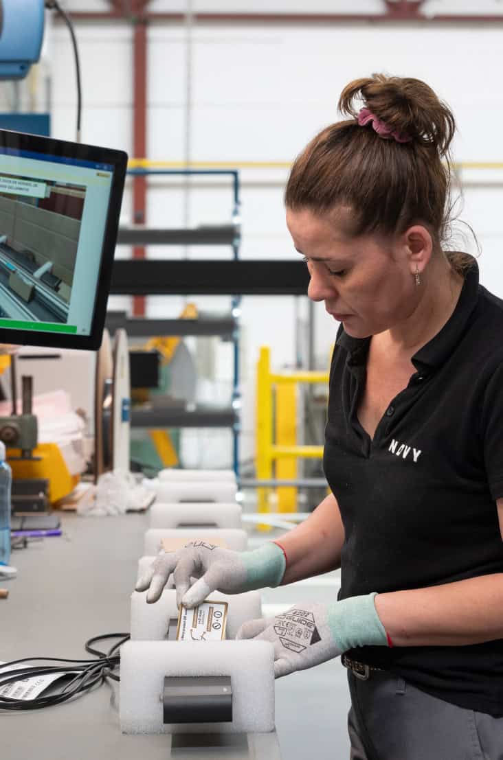 A woman in a black polo shirt and gloves is working at a workstation. She is holding a rectangular object and inspecting it. Behind her, there is a computer monitor displaying some images, and various equipment and materials are visible in the background.